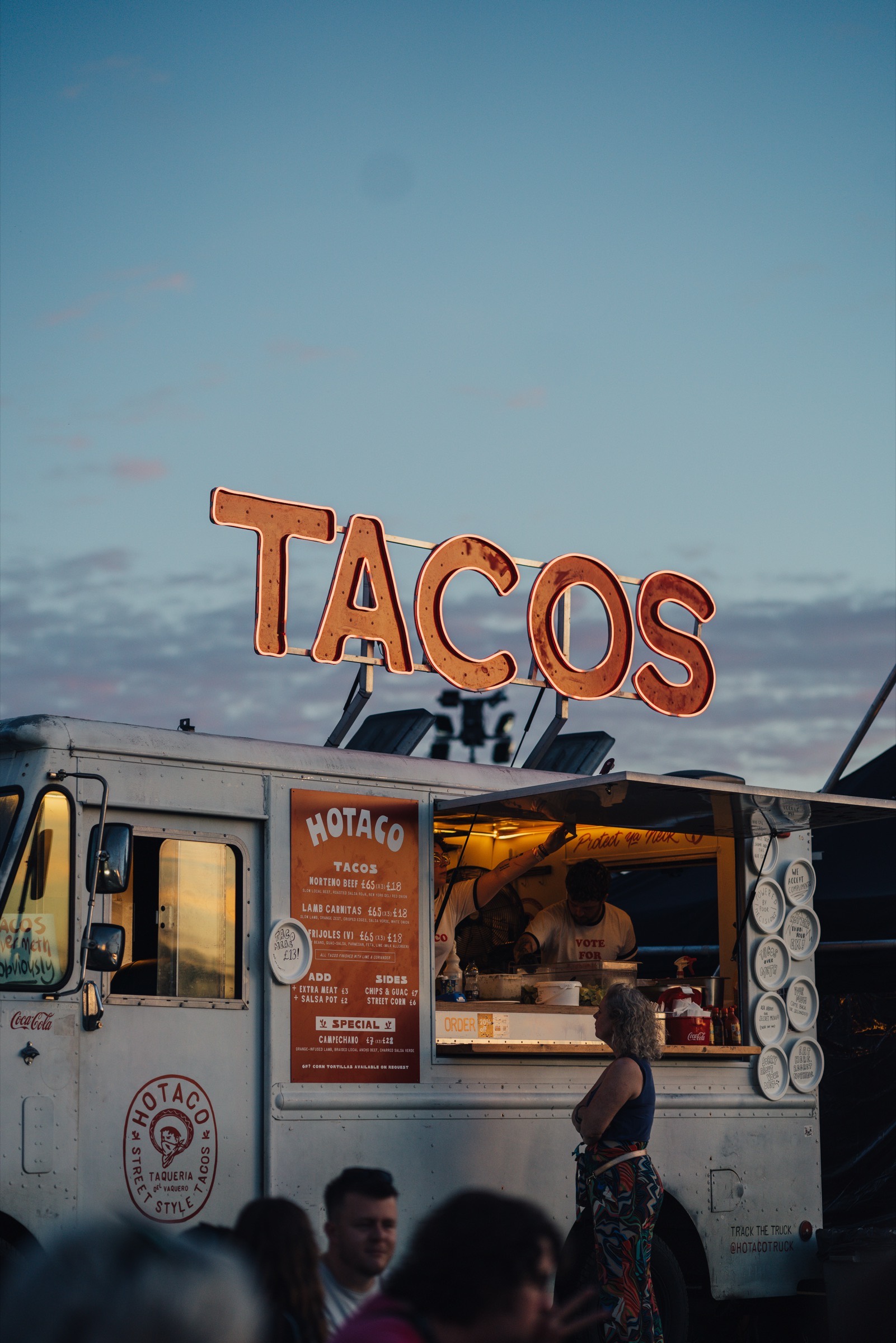 The step van at dusk, the TACOS sign above it and a customer at the hatch.