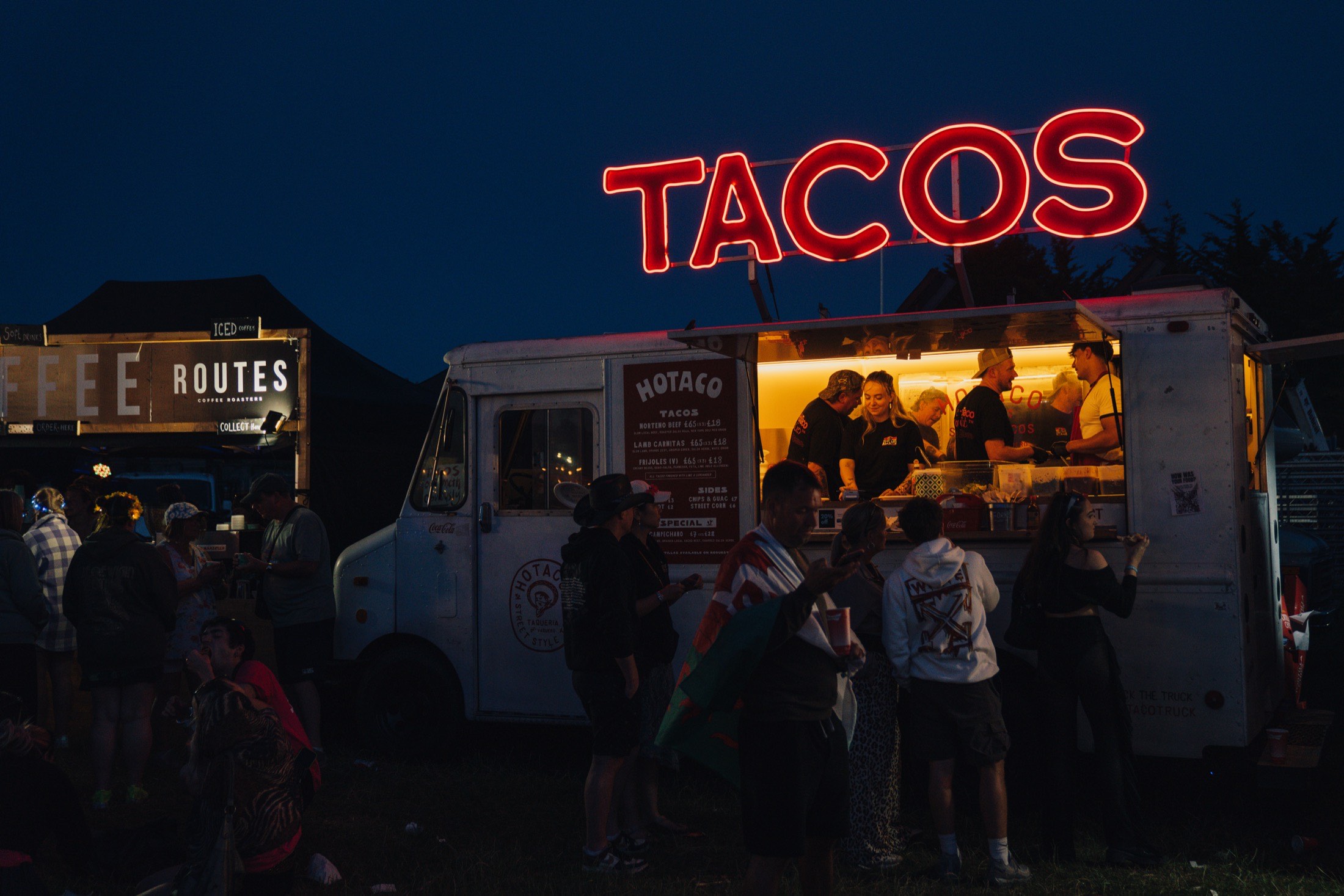 The HoTaco step van at night, the neon TACOS sign lit red above it, the hatch glowing and a queue at the window.