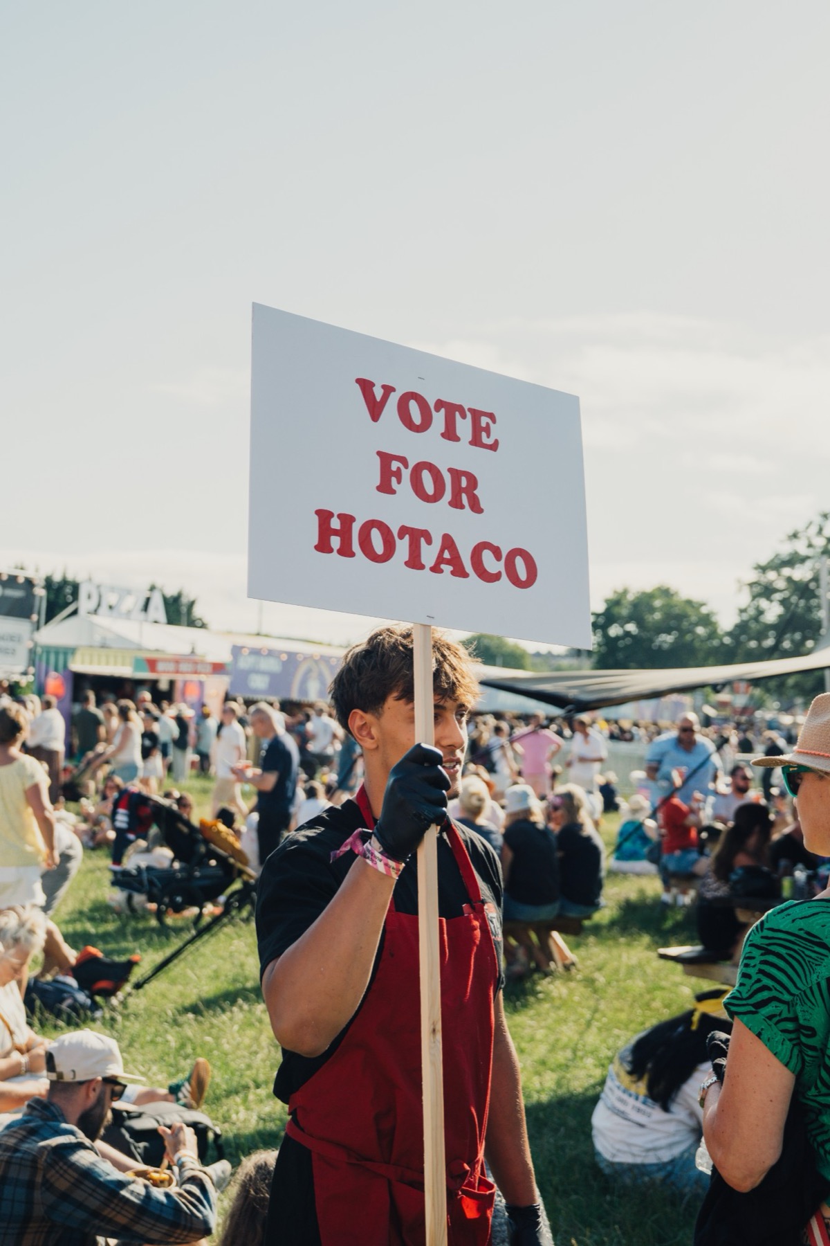 A member of the HoTaco crew holding a Vote For HoTaco placard in a busy festival crowd.