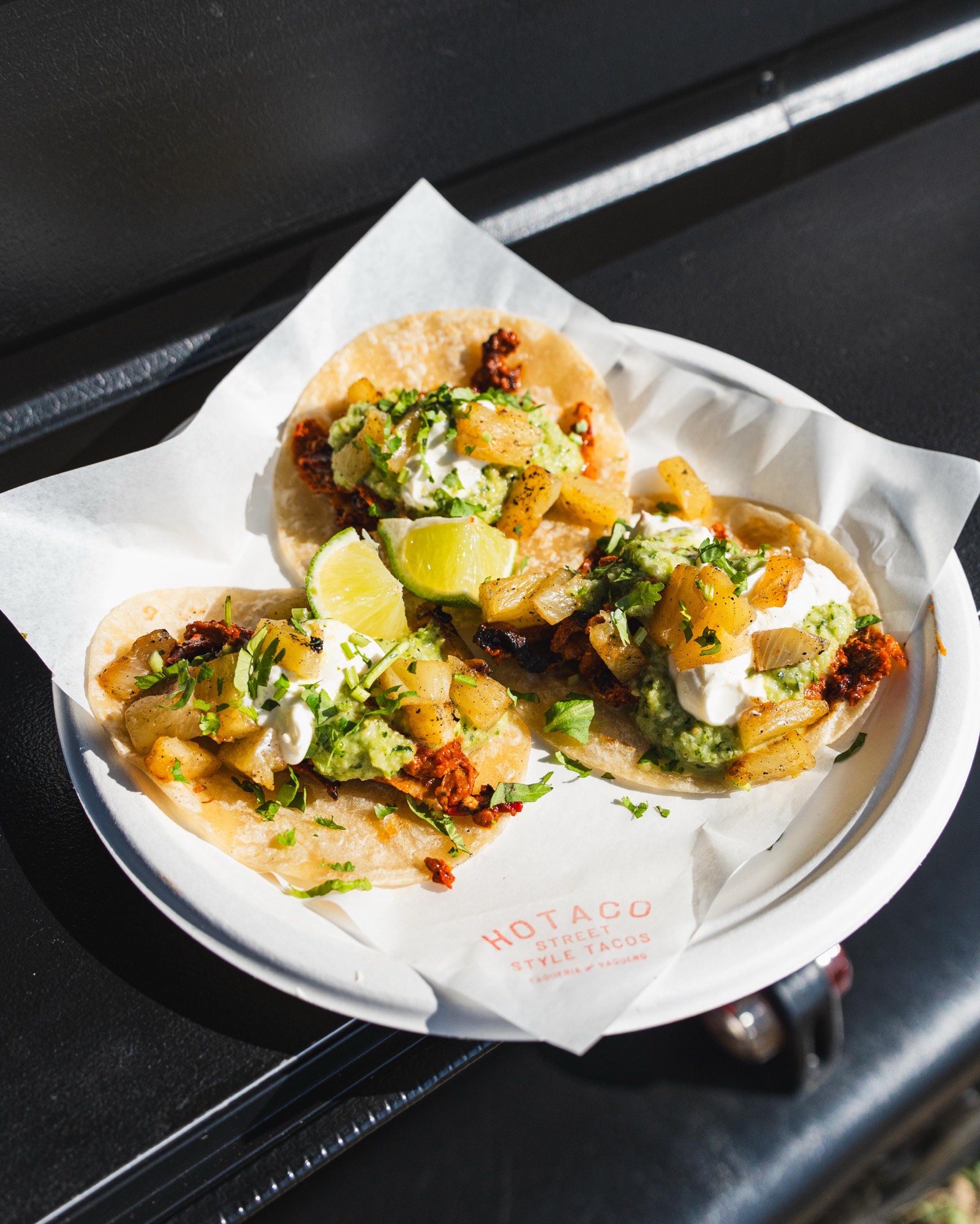 Three al pastor tacos on HoTaco greaseproof paper: pork, charred pineapple, guac salsa, sour cream, coriander and lime.
